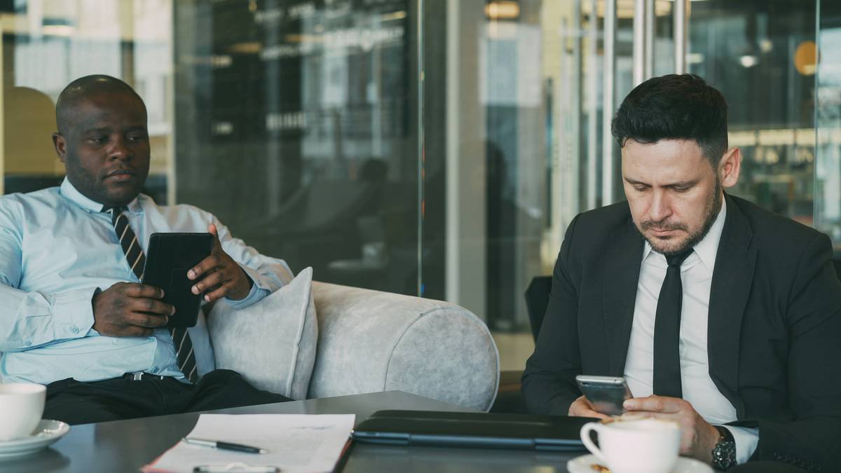 Two multi-ethnic businessmen in formal clothes sitting and using their smartphone and tablet in glassy cafe during lunch time indoors