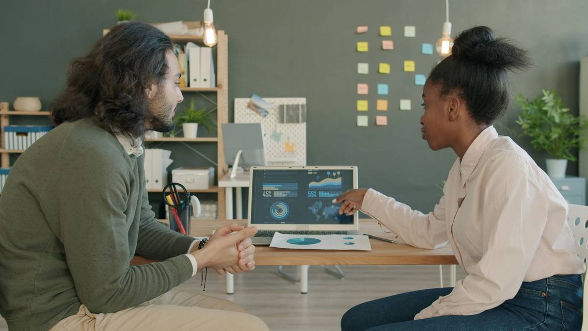 Colleagues mixed race man and Afro-American woman are talking and using laptop in office discussing business ideas. People, workplace and job concept.