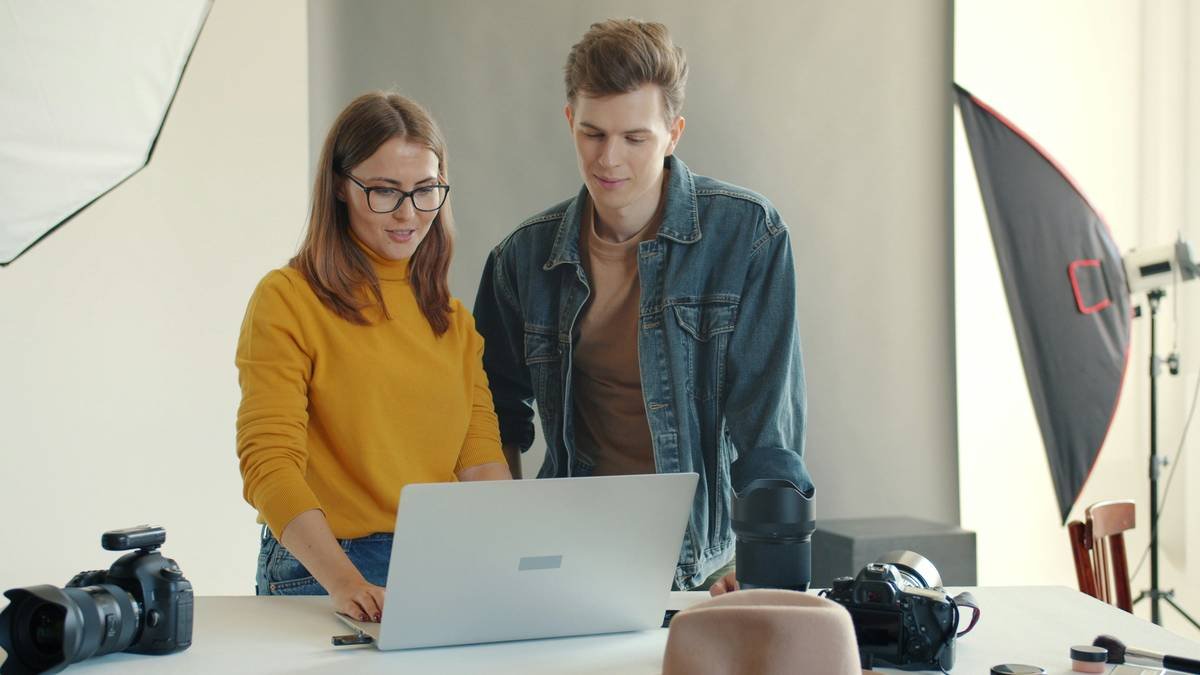 Happy young man and woman are using laptop in studio watching photos after photoshoot talking discussing pictures. People and communication concept.